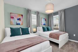 Experience a bright and airy bedroom with two cozy beds adorned with plush teal pillows and soft throws, vibrant artwork for a pop of color, and large windows that bathe the room in natural light, ensuring a refreshing stay.