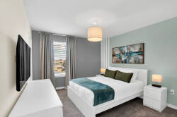 Experience a serene bedroom featuring calming gray and green tones, with a cozy bed dressed in crisp white linens. Natural light pours in through a large window, accentuating a modern art piece that brings character to the room.