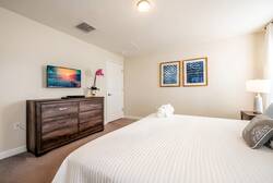 This cozy bedroom boasts a plush white bedspread for relaxation, with soft natural light streaming through sheer curtains. A stylish wooden dresser and colorful artwork enhance the charm, promising a restful stay.