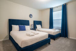 This cozy bedroom offers two plush beds with blue headboards and soft light gray carpeting, creating a warm atmosphere. Natural light streams through the window, making it an inviting space for restful nights.
