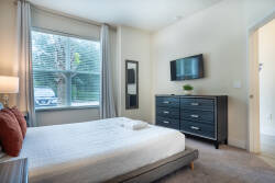 This bright and airy bedroom offers a plush queen bed and a stylish black dresser that harmonizes with the soft, neutral walls. Enjoy the warm ambiance created by natural light streaming in through the large window.