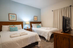 This cozy bedroom offers two comfy beds with soft white linens and decorative pillows, creating a welcoming vibe. A warm wooden dresser and side table enhance the light blue walls, while soft lighting adds a soothing touch.