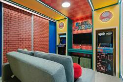 Discover a vibrant gaming nook featuring cheerful yellow walls and classic brick accents, ideal for relaxation and fun. The cozy gray sofa invites you to unwind, while a retro arcade game and colorful decor ensure endless.