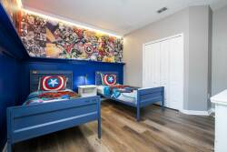 Bright blue walls set a playful tone in this cozy bedroom, featuring two charming twin beds adorned with superhero-themed bedding. A vibrant mural adds character, while warm wood flooring enhances the inviting atmosphere.