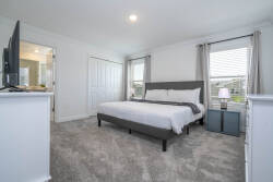 This bright and spacious bedroom, with its plush carpet and stylish gray bedframe, invites relaxation. Enjoy a wall-mounted TV, a cozy bedside lamp, and soft curtains that frame a refreshing view for your comfort.