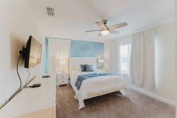 Experience tranquility in this cozy bedroom with soft beige carpeting and a calming blue accent wall. Stylish white furniture, including a plush bed and sleek bedside tables, enhances the serene ambiance created by natural light.