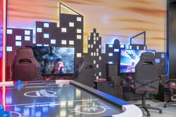 Experience a vibrant gaming haven with sleek black chairs, a dynamic air hockey table, and large screens showcasing thrilling visuals against a city skyline. Colorful lighting sets the perfect mood for unforgettable game nights.
