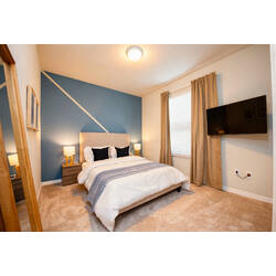 Rest in a cozy bedroom with a white bed, blue accent wall, warm bedside lamps, wall-mounted TV, soft curtains, and neutral carpet for vacation comfort.