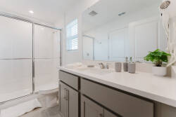 This bright and airy bathroom boasts a sleek dual-sink vanity with a white countertop and stylish gray cabinetry. Enjoy refreshing moments in the glass shower enclosure, enhanced by a touch of greenery for a vibrant accent.