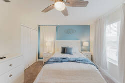 Experience a bright and airy bedroom with a calming blue accent wall and crisp white bedding. Enjoy the warmth of plush carpet underfoot, complemented by a ceiling fan and natural light for a refreshing, restful atmosphere.