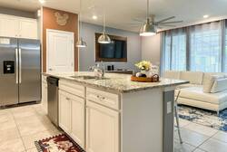 Cook and gather in a bright kitchen with white cabinets, granite island, stainless steel appliances, pendant lights, and an open view to the living room, perfect for family vacation moments.