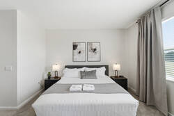Rest in a bright and cozy bedroom with a comfortable bed, soft white bedding, gray accents, bedside lamps, abstract wall art, carpeted flooring, and a large window with natural light.
