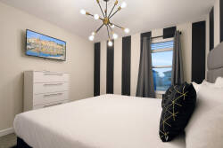 Experience a vibrant bedroom with bold black-and-white striped walls that highlight a plush bed topped with a stylish geometric pillow, all illuminated by a modern chandelier and complemented by a sleek dresser and a lovely view.