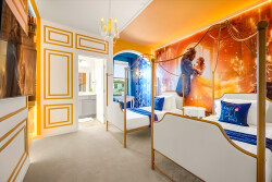 Experience a whimsical retreat with two charming beds framed in gold, bathed in soft lighting. The vibrant orange and blue hues, along with a captivating wall mural, create a playful atmosphere ideal for relaxation and enjoyment.