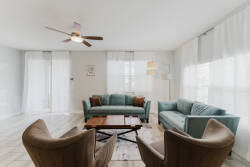 Experience the bright and airy living area with plush teal sofas and warm wood accents. Large windows with sheer curtains let in soft light, making it a perfect space for relaxation and enjoyable conversations.
