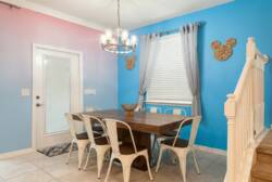 Gather in a bright dining area with a wooden table, white chairs, blue accent walls, soft curtains, modern chandelier, and staircase nearby, perfect for family meals during vacation.
