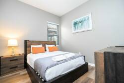 This cozy bedroom boasts a stylish wooden bed with crisp white linens and vibrant orange accents, creating a cheerful atmosphere. A warm bedside lamp glows softly, and a large window invites natural light and refreshing views.