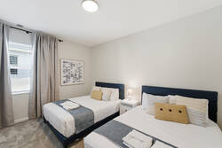 Rest in a bright bedroom with two comfortable beds, soft white bedding, blue headboards, gray curtains, bedside lamp, wall art, carpeted floor, and warm neutral decor.