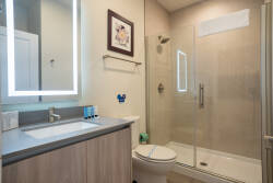Experience a bright, airy bathroom with a sleek glass shower and stylish floating vanity featuring elegant gray countertops. Warm lighting and tasteful floral artwork enhance the soothing atmosphere for your daily routine.