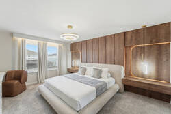 Rest in a bright modern bedroom with a cozy white bed, gray accent throw, warm wood feature wall, soft pendant lights, elegant ceiling fixture, white curtains, and a comfy chair for vacation comfort.