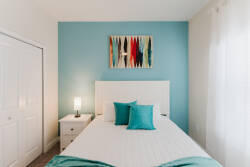This cozy bedroom features a calming blue accent wall, crisp white bedding, and vibrant teal pillows. Natural light streams through sheer curtains, creating an inviting atmosphere that highlights the stylish wall art above the.