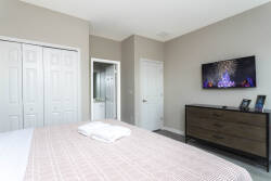 This cozy bedroom boasts a plush light-hued bed with a soft quilt, a dark wood dresser, and a sleek flat-screen TV for your entertainment. The neutral tones and natural light create a warm atmosphere, perfect for relaxation.