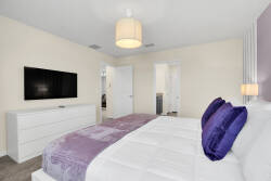 Welcome to a chic bedroom with a plush lavender-accented bed for ultimate relaxation. The sleek white dresser and modern lighting create an inviting space, while the wall-mounted TV adds to your comfort for a restful night's.