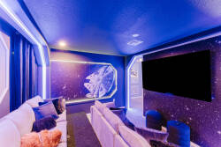Experience a cozy entertainment room with plush white seating and colorful pillows. The vibrant blue and purple lighting enhances the atmosphere, while a large screen is ready for your memorable movie nights.