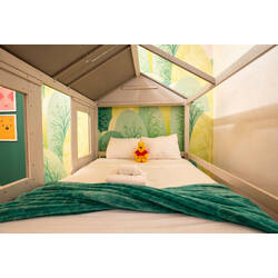 Sleep in a cozy house-style kids bed with soft bedding, green blanket, and a cheerful forest mural. Warm lighting, playful details, and a sweet teddy bear create a calm and fun space for little guests during vacation.