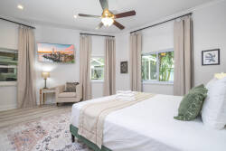 Relax in this beautifully decorated bedroom filled with natural light, featuring a plush white bed with soft linens, calming neutral tones, a cozy armchair, and vibrant artwork that adds a delightful touch of color.