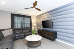 Experience a cozy living area with soft gray tones and a striking striped wall, creating a warm atmosphere. Relax on the plush sectional, complemented by a sleek coffee table and modern fan that enhance the stylish appeal.