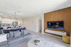 Relax in a bright open living area with cozy gray seating, a wall-mounted TV, warm wood accent wall, dining space, modern kitchen access, and soft decor for comfortable family time.