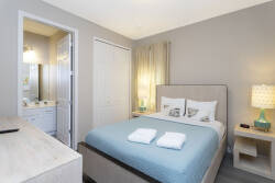 Enjoy restful nights in this serene bedroom, featuring a cozy blue bedspread, soft lamp lighting, and a convenient en-suite bathroom nearby. The calming neutral tones create a perfect atmosphere for your relaxing getaway.