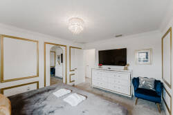 Welcome to this beautifully styled bedroom, featuring a plush bed under a sparkling chandelier. Elegant gold accents brighten the crisp white walls, while a cozy armchair and modern dresser add to the inviting ambiance.