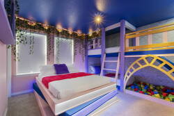 This bright and playful children's room features a cozy double bed with vibrant bedding, a stylish bunk bed, and a colorful ball pit. The rich blue ceiling and leafy accents create a whimsical atmosphere for your little ones.