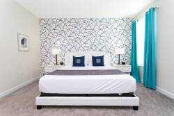 Bright turquoise curtains frame large windows, letting in natural light that highlights the modern geometric wallpaper behind a cozy, neatly made bed.