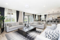 Experience a sunny living area with large windows that fill the space with natural light, showcasing soft gray sofas with stylish pillows. The modern coffee table and chic rug add a cozy, sophisticated touch, ideal for relaxation.