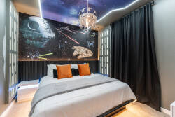Experience a unique bedroom with whimsical decor and modern comfort, featuring a galaxy mural and soft lighting. Cozy bedding with vibrant orange pillows invites relaxation, all set on warm, sleek wooden floors for a perfect.