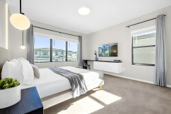 This stylish bedroom, filled with natural light, offers a plush bed with crisp white linens and a cozy throw. Its minimalist decor and modern pendant lighting create a serene atmosphere ideal for your relaxation.