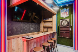 Experience the stylish game room with a beautifully crafted wooden bar and sleek stools, perfect for enjoying drinks while watching sports on the large TV. The vibrant neon lighting adds a fun touch to this inviting space.