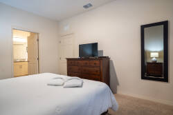 Experience a cozy bedroom with a plush bed dressed in crisp white linens, a dark wood dresser, and a large mirror. Soft, neutral tones create an inviting ambiance, with natural light filtering in from the bathroom door.