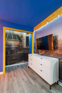 Bright blue walls and vibrant yellow accents create a playful atmosphere in this stylish room. The sleek white dresser complements the modern decor, while the wall art hints at culinary adventures just beyond the door.