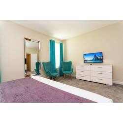 Rest in a bright modern bedroom with a comfortable bed, white bedding, blue accent pillows, geometric wall design, teal curtains, bedside lamps, soft carpet, and a stylish accent chair.