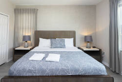 Welcome to a cozy bedroom perfect for relaxation, with a plush bed dressed in a stylish gray and white blanket. Soft bedside lamp lighting creates a warm atmosphere, beautifully complemented by elegant curtains framing the window.