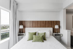 This bright and airy bedroom features a plush white bed with textured green pillows, a warm wood headboard, and soft gray tones, while large windows fill the space with natural light for a tranquil atmosphere.