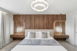 Rest in a bright modern bedroom with a cozy white bed, gray accent throw, warm wood feature wall, soft pendant lights, elegant ceiling fixture, white curtains, and a comfy chair for vacation comfort.