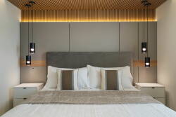 Enjoy peaceful nights in this chic bedroom with a plush gray headboard and cozy linens. The sleek bedside lamps cast a warm glow, while wood paneling enhances the inviting and sophisticated ambiance.