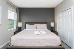 Awake to gentle natural light in this cozy bedroom, featuring a plush bed with soft linens and sleek bedside lamps. The calming grey accent wall creates a tranquil atmosphere, ensuring restful nights for your stay.
