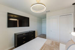 This cozy bedroom features a sleek black dresser and a wall-mounted TV for your entertainment. A contemporary light fixture casts a warm glow, enhancing the inviting atmosphere of the soft beige textiles and neutral walls.