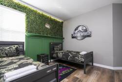 This playful bedroom features two stylish black beds with colorful dinosaur-themed bedding and warm wooden floors. A vibrant green accent wall with lush plants brings fun, while natural light creates a bright, cheerful atmosphere.